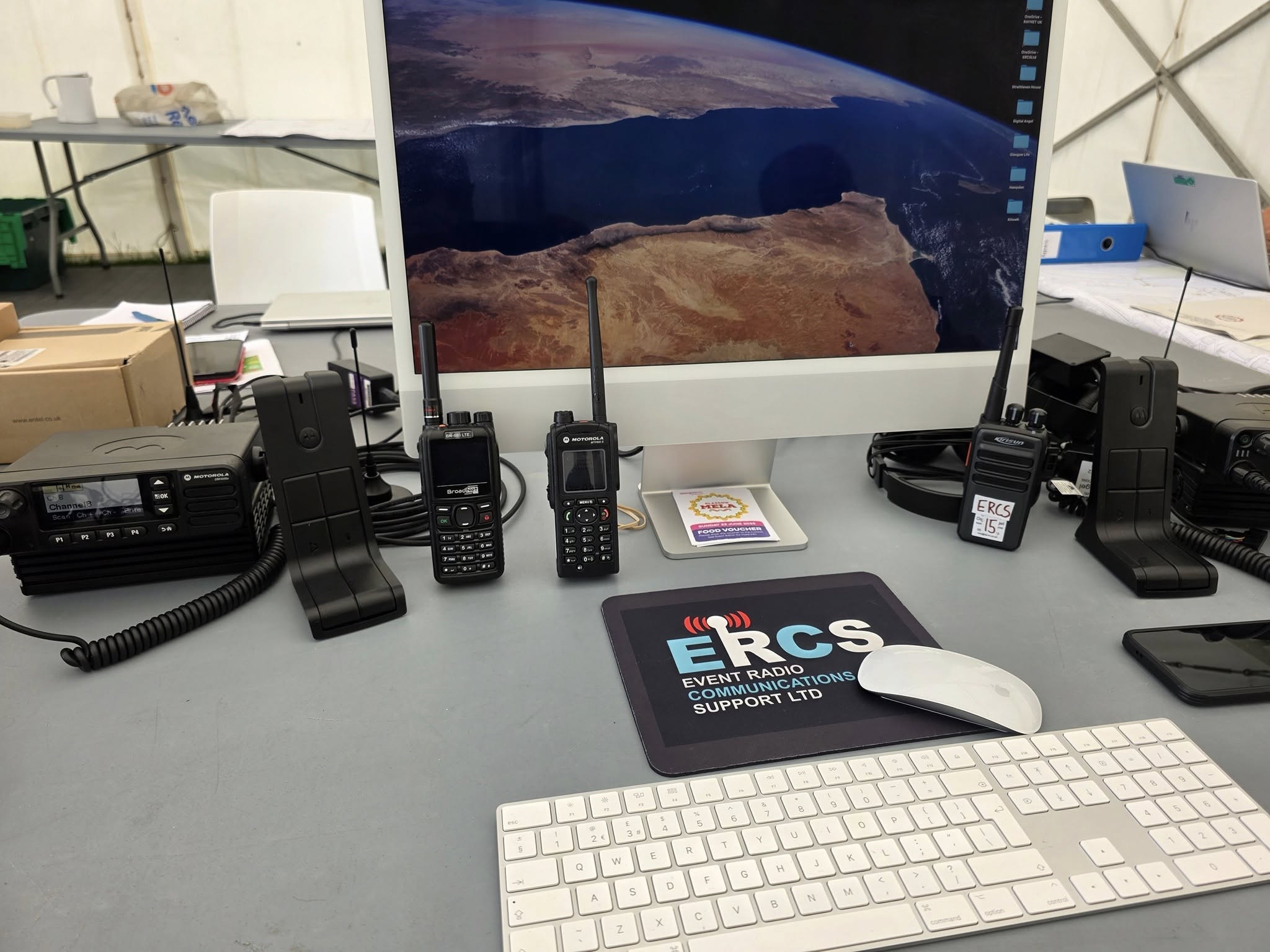 ERCS control-desk equipment — mobile base radio, Motorola digital handsets, BroadNet LTE terminal and ERCS-branded mousepad at the Glasgow Mela 2025 set up.